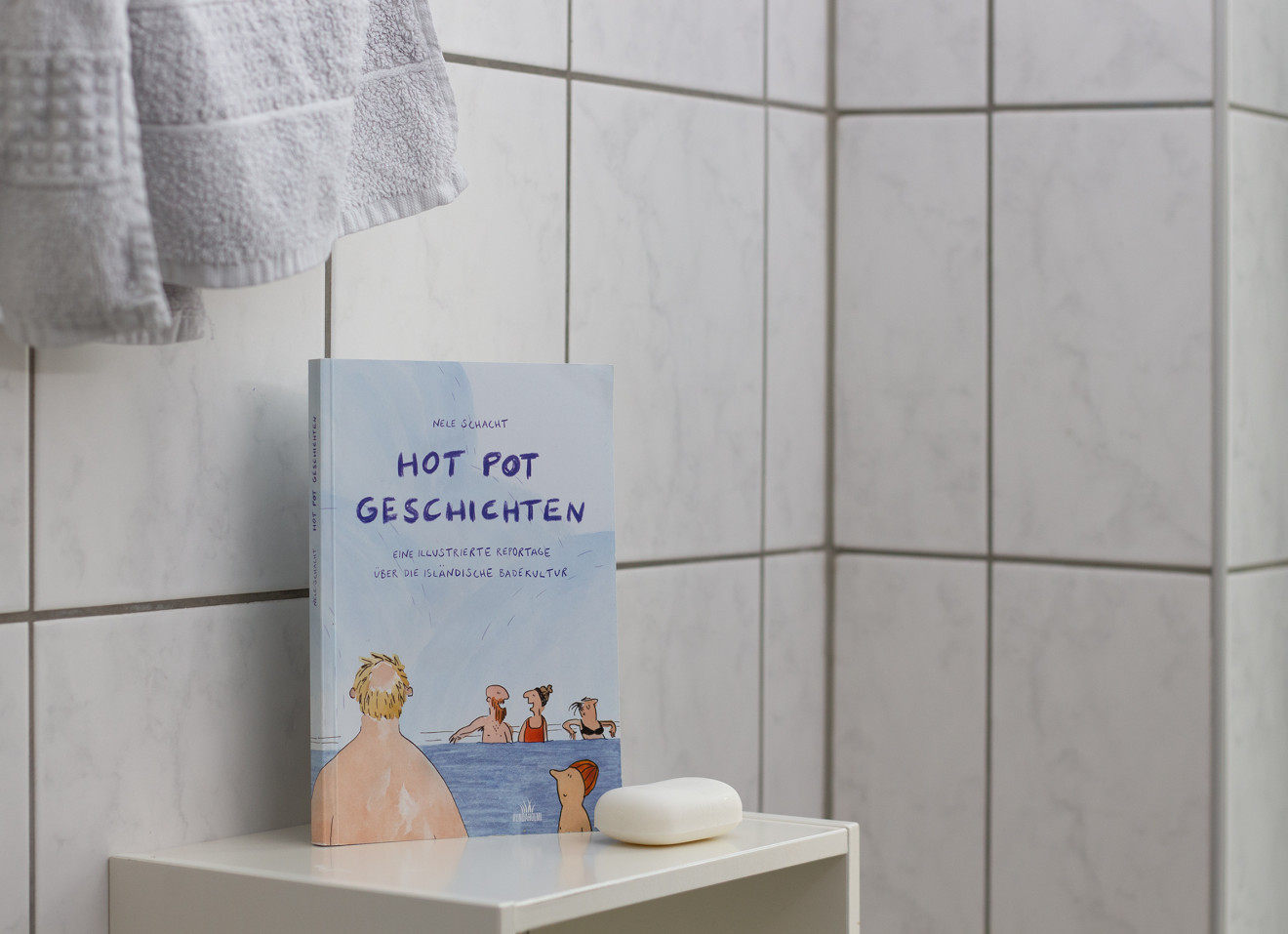 Das Buch steht vor einer gefliesten Badezimmerwand. Auf dem Buchtitel befindet sich eine Zeichnung von mehreren Menschen, die in einem Thermalbad sitzen und sich unterhalten.
