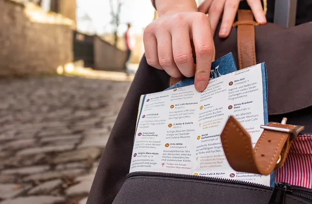 Eine Hand holt den Stadtplan aus der Außentasche eines Rucksacks. Im Hintergrund sieht man eine sonnige Straße.