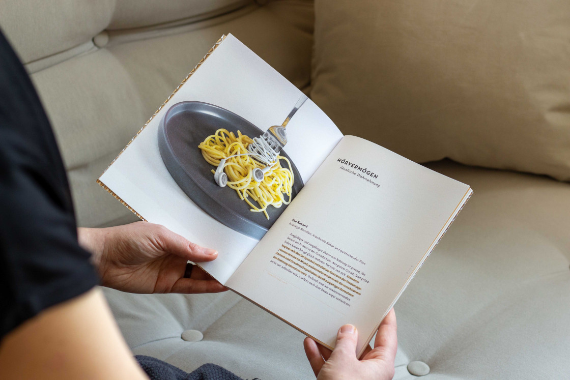 Das aufgeschlagene Heft Ohrenschmaus wird in Händen gehalten. Das Foto im Heft zeigt einen Teller Spaghettis. Darin steckt eine Gabel mit am Kabel aufgedrehten Kopfhörern.