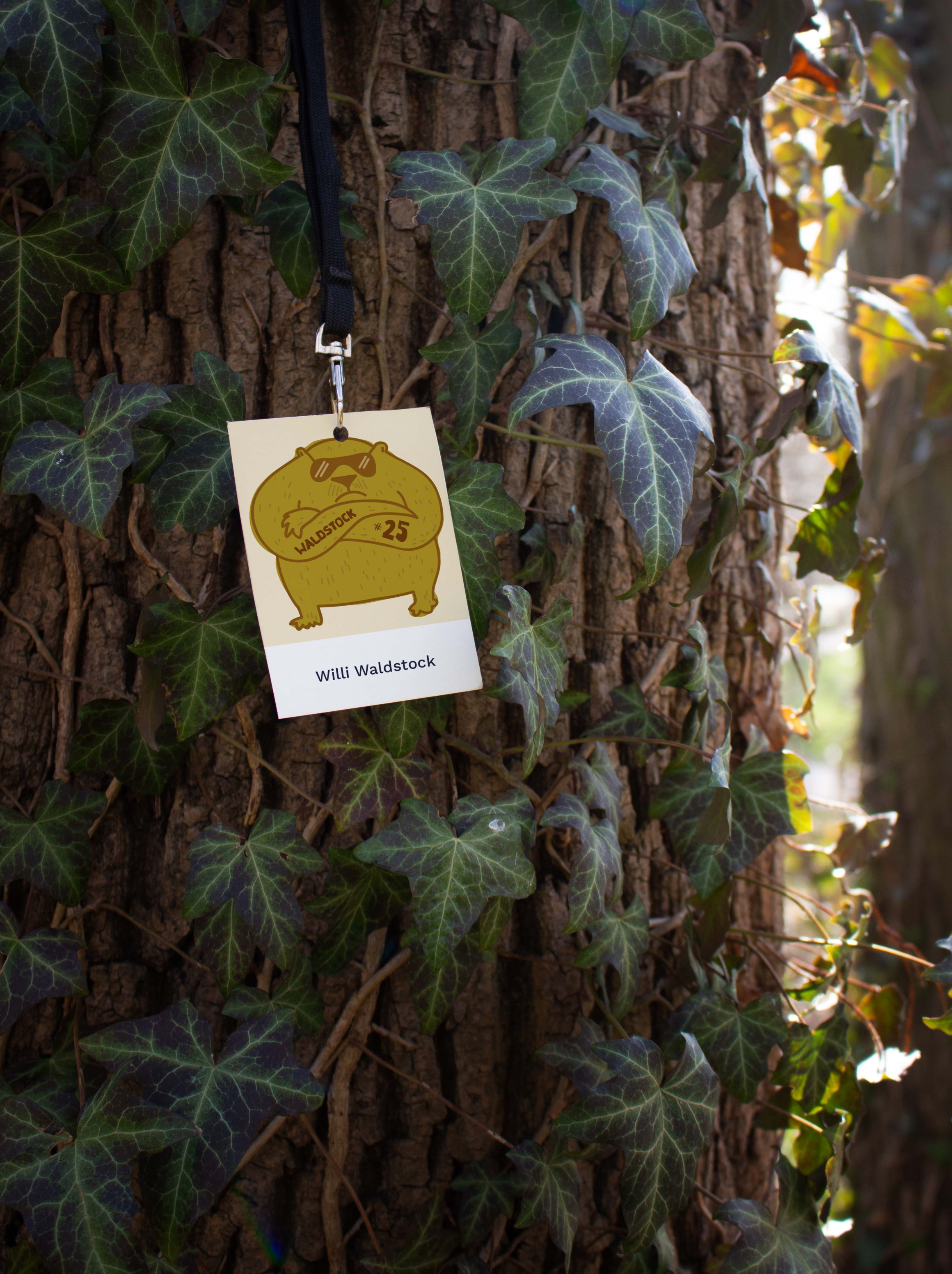 Backstage-Pass hängt an einem Baumstamm, der mit Efeu bewachsen ist. Auf dem Pass ist die Zeichnung eines dicken Bären, der das Waldstock-Logo auf den Unterarm tätowiert hat.