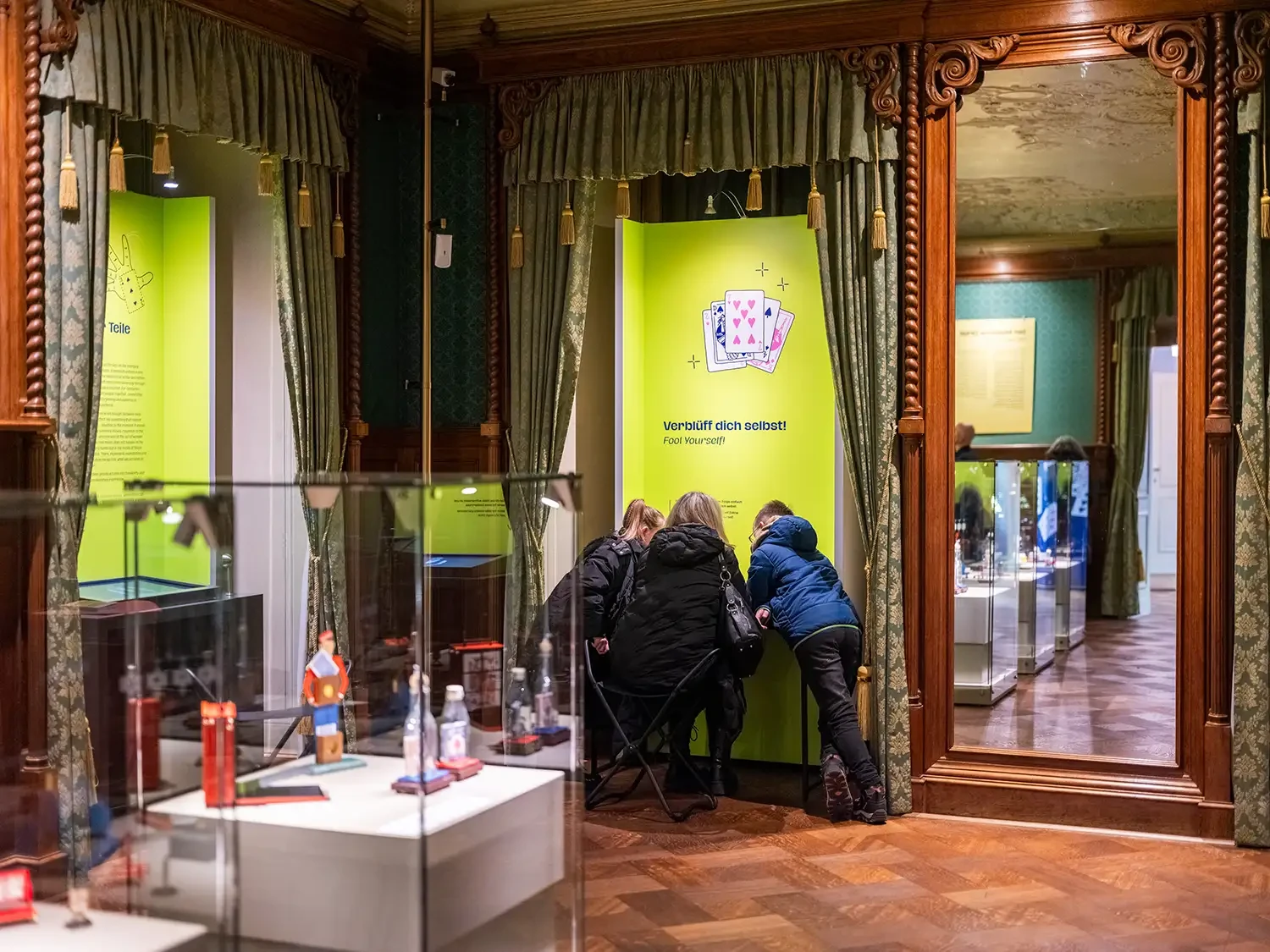 Detail im dritten Ausstellungsraum. Vor einer Texttafel stecken drei Personen die Köpfe zusammen, um einen Kartentrick zu lernen.