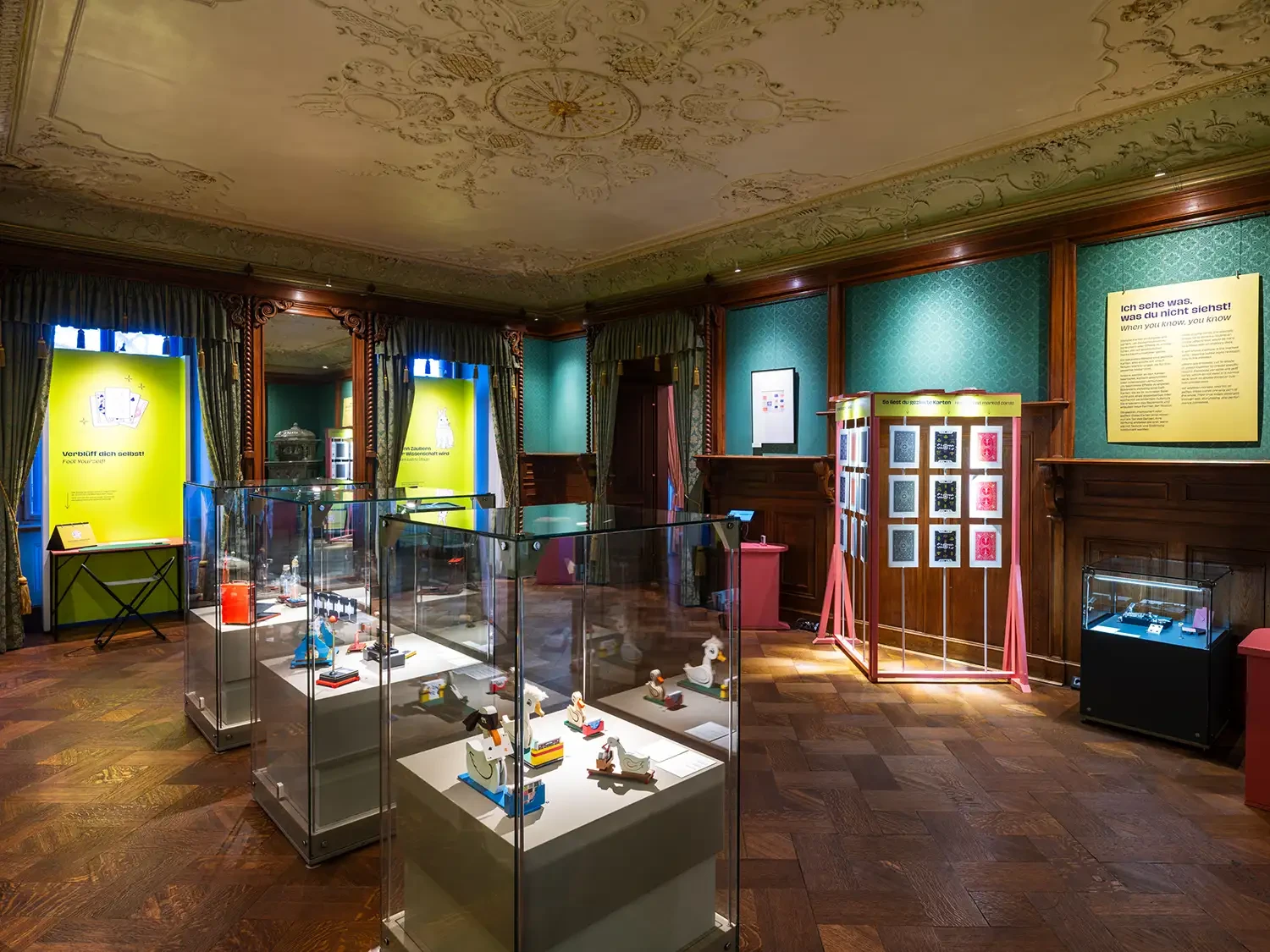 Blick in den dritten Ausstellungsraum. In den Fensternischen befinden sich Texttafeln und in der Mitte des Raumes Vitrinen mit Ausstellungsstücken.